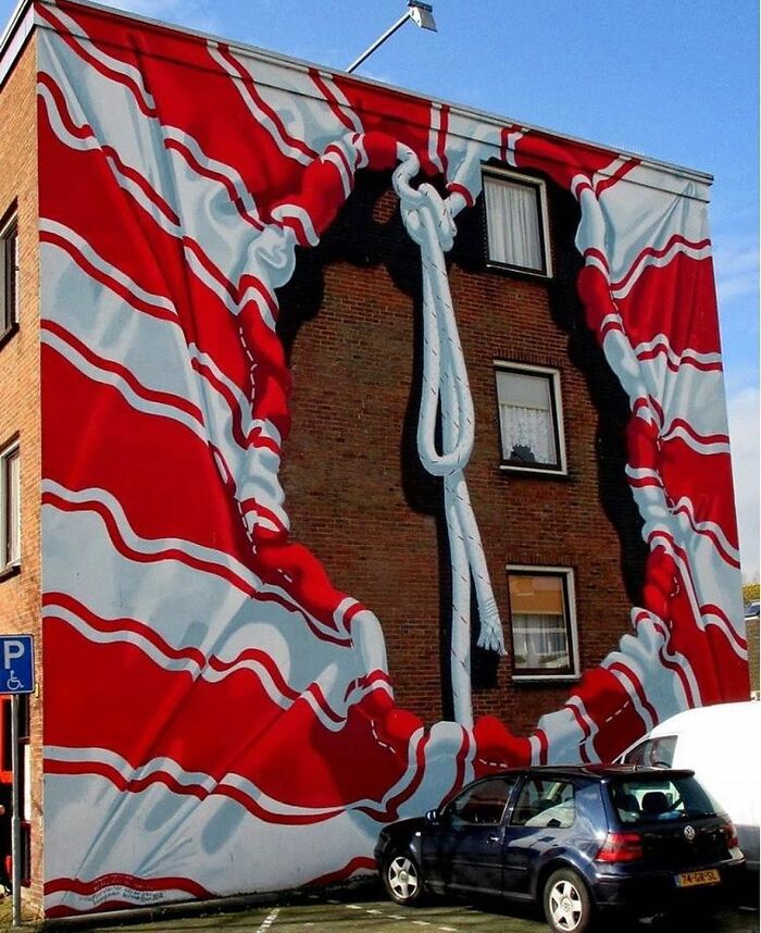 Optical illusion street art on a brick building, depicting red and white fabric pulled back with a knotted rope.