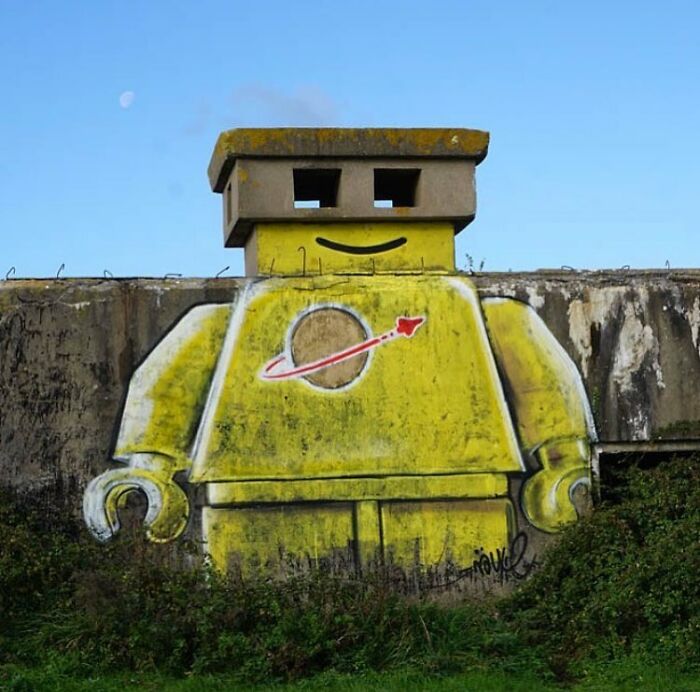 Giant yellow LEGO character street art painted on a concrete structure, with a small moon in the sky.