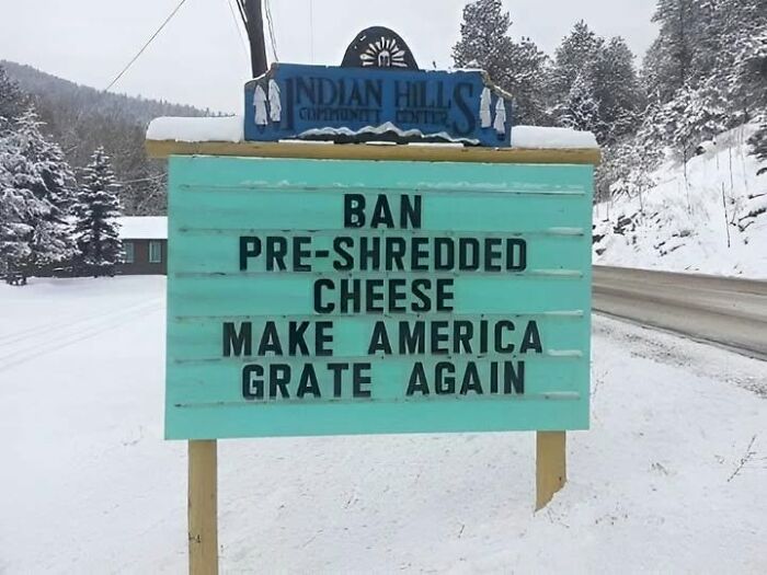 A funny sign in a snowy landscape reads BAN PRE-SHREDDED CHEESE MAKE AMERICA GRATE AGAIN. This silly sign adds humor to a winter scene.