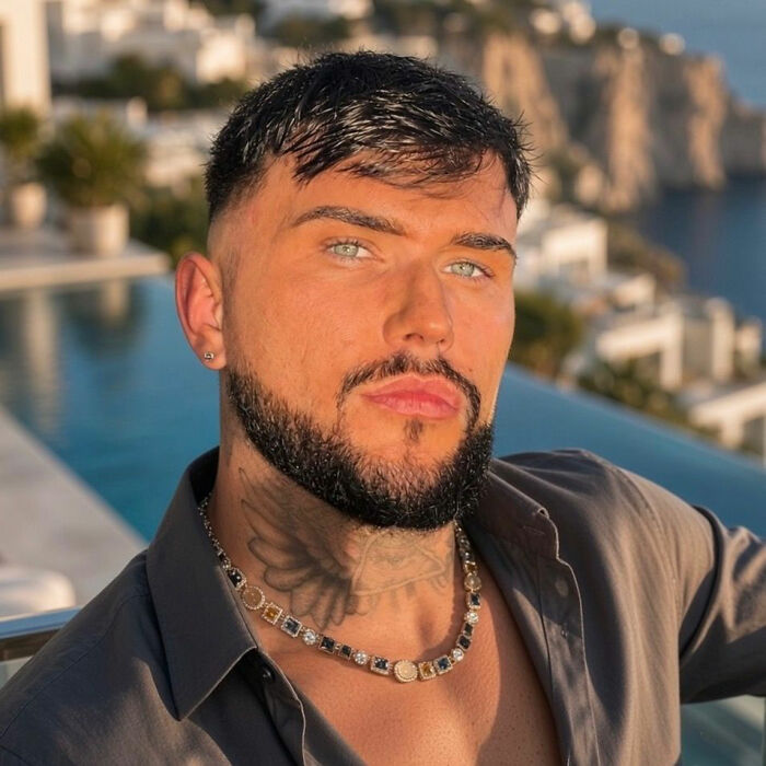 A handsome male hairdresser with dark hair and striking blue eyes, posing in front of an ocean view.