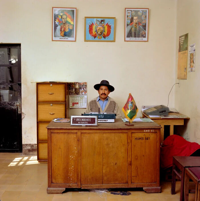 A photographer's project presenting a man in a hat at a wooden desk, documenting the people and offices behind bureaucracy worldwide.