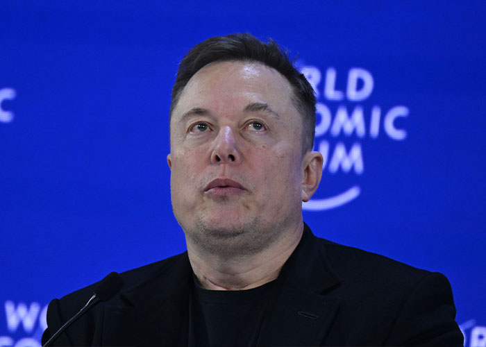Close-up of Elon Musk speaking on a panel in front of a blue background