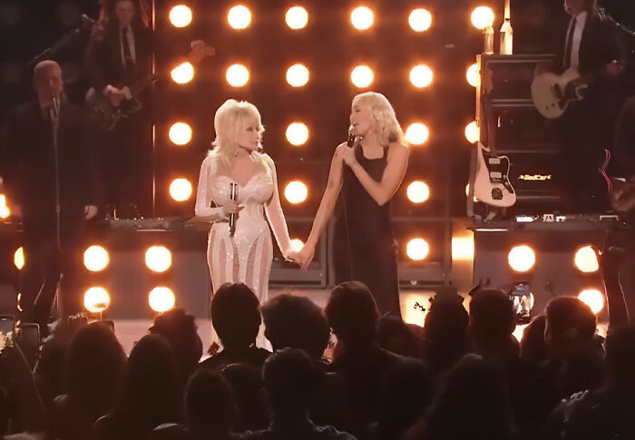 Miley Cyrus and Dolly Parton holding hands and singing on stage at a concert, surrounded by bright lights and a band.