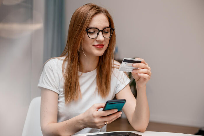 A young woman in glasses holding a credit card and smartphone, depicting dumb ways people ruin their lives via financial irresponsibility.