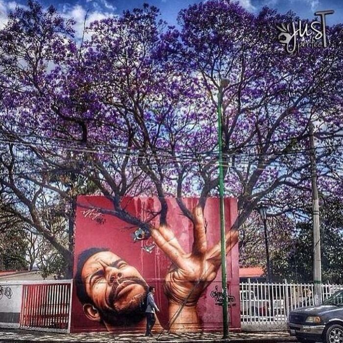 A vibrant street art mural of a man's face and a hand forming a peace sign on a red wall, with purple jacaranda trees above.