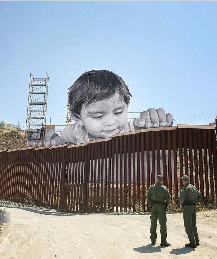 Giant baby street art mural peeking over a border fence, with two guards walking below, a stunning example of street art.