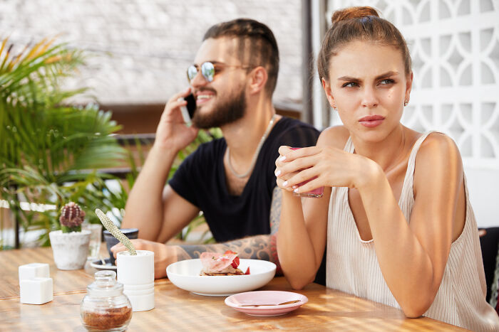 A woman looking annoyed while a man talks on the phone, illustrating dumb ways people ruin their lives in relationships.