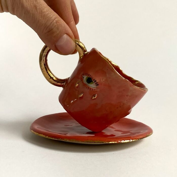 A small pink ceramic sculpture cup with a painted eye and gold handle, resting on a matching saucer.