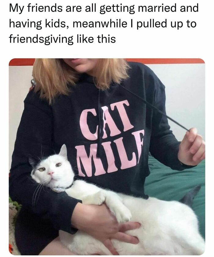A woman wearing a CAT M**F hoodie holds a fluffy white cat with black spots on its ears and tail. This cat meme is impossible not to laugh at.