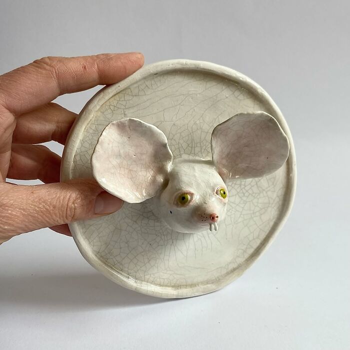 A hand holding a round ceramic sculpture plate with a quirky white mouse head on it.