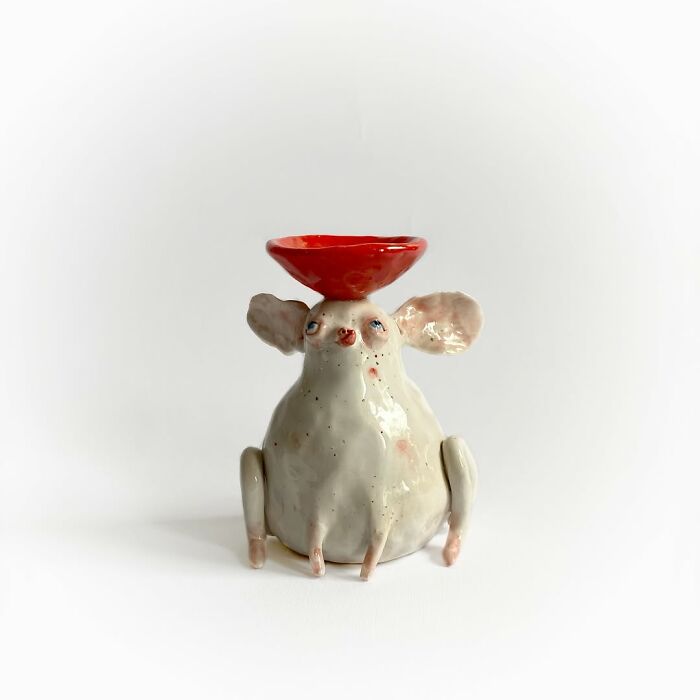 Weird ceramic sculpture of a whimsical white creature holding a small red bowl on its head.