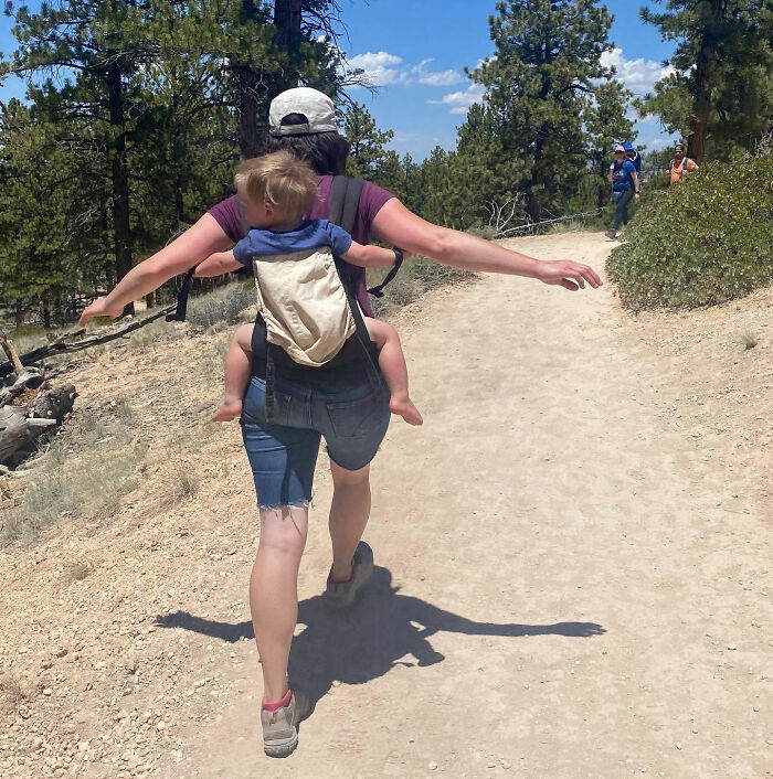 Mom carrying her son in a baby carrier on a dirt path with trees, showing a contrasting protective act. Mom carrying her son in a baby carrier on a dirt path with trees, showing a contrasting protective act.