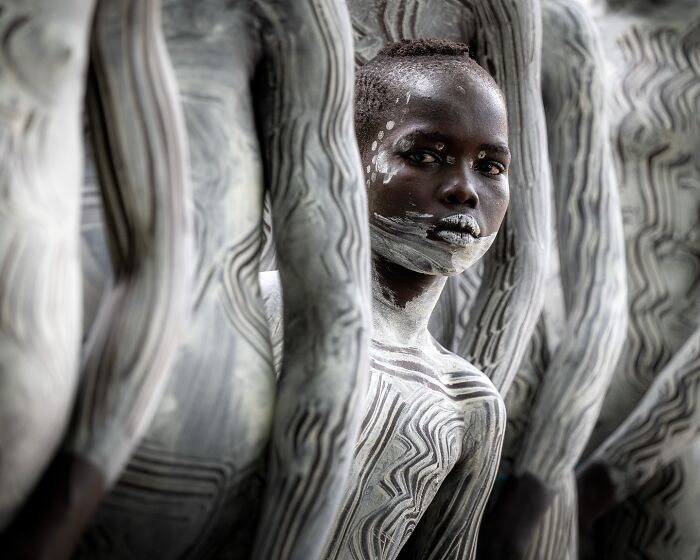 A young person with dark skin and white body paint looking directly at the camera, a remarkable person in travel photos.