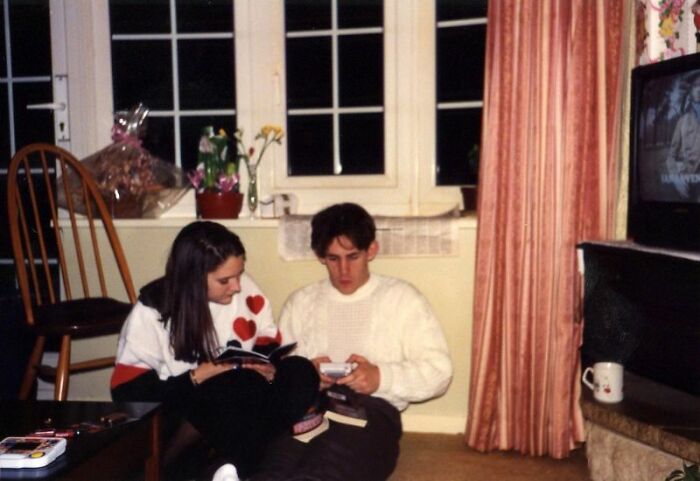 A couple sitting indoors, one holding a handheld 80s retro tech device, a glimpse of glorious weirdness.