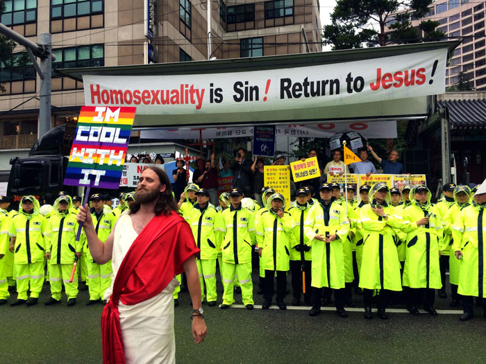 A person dressed as Jesus holding a rainbow flag, standing in front of a protest banner that reads 'Homosexuality is Sin! Return to Jesus!'