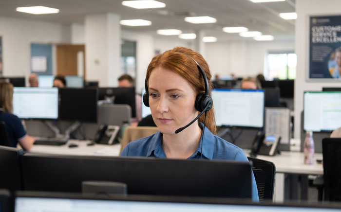 A focused worker with a headset works at a computer in a busy, open-plan office environment.