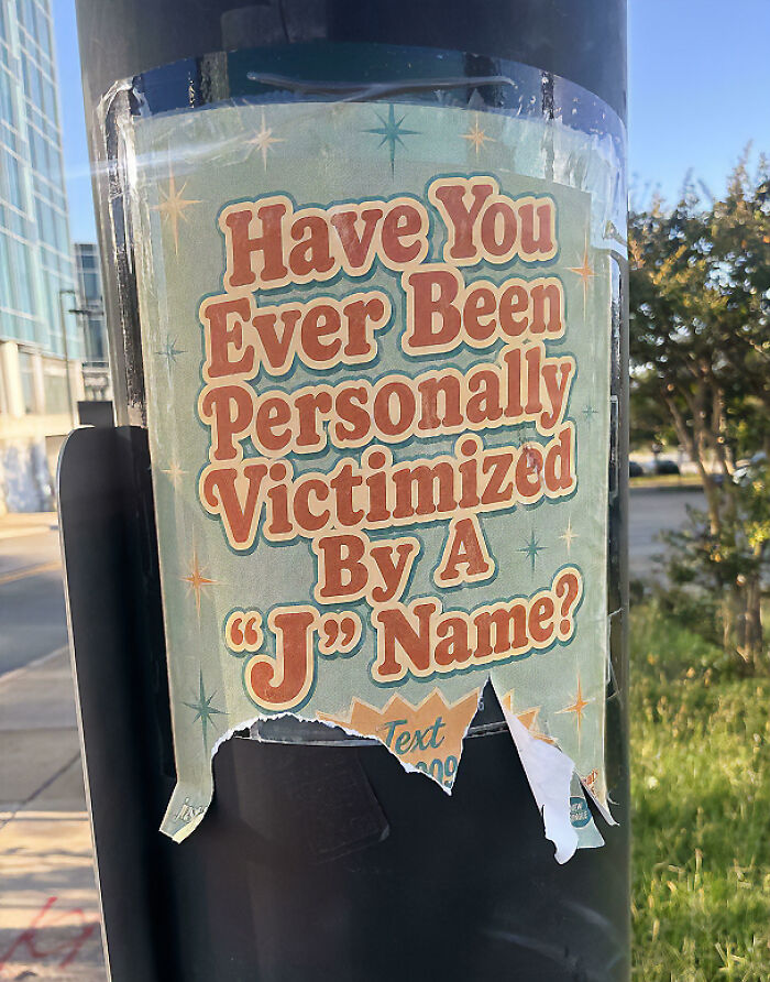 A chaotic sign on a pole asks 'Have You Ever Been Personally Victimised By A “J” Name?', making people question reality.