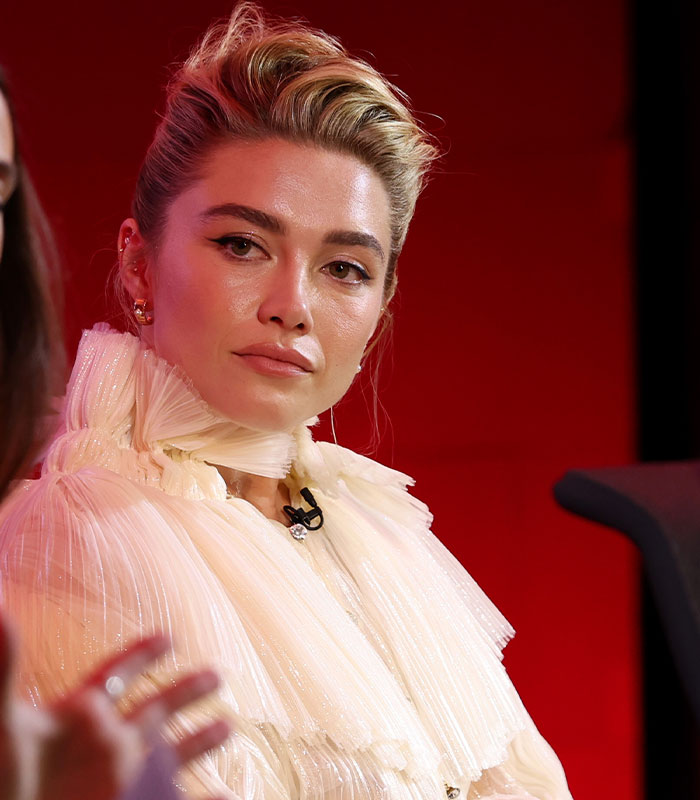 Florence Pugh wearing a high-collared white ruffled top with a sleek updo hairstyle. Florence Pugh wearing a high-collared white ruffled top with a sleek updo hairstyle.