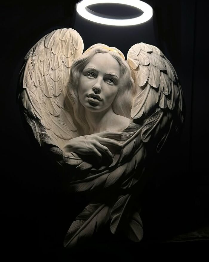 A lifelike wood sculpture of a female angel with wings, illuminated under a ring light by a Polish artist