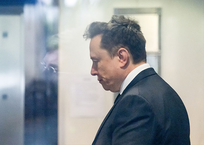 Side profile of Elon Musk looking down in a dark suit, amidst rumors of a Spaceport visit and scoliosis.