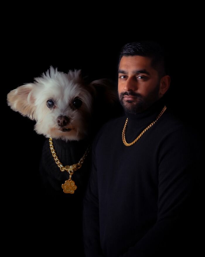 Quirky portrait of a man and his small white dog, both wearing black turtlenecks and gold chains, against a dark background.