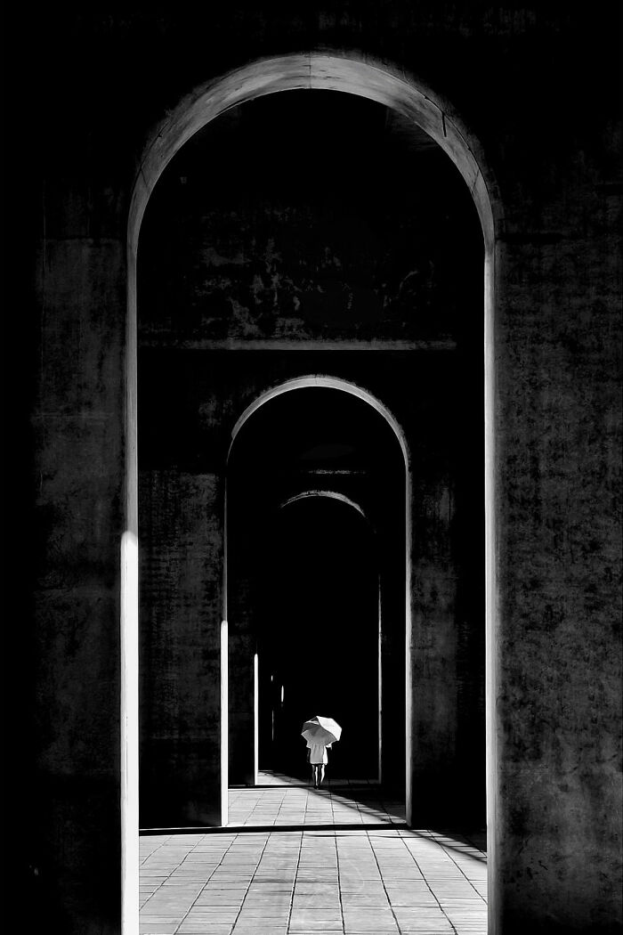 A photographer's minimalist architecture scene with a person holding an umbrella walking through a series of dark arches.