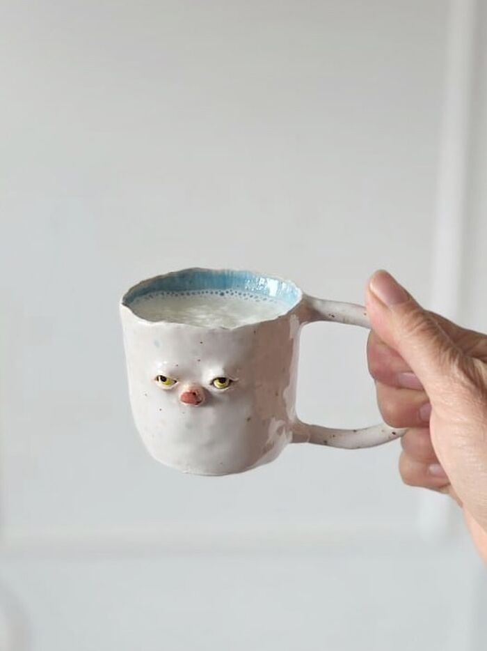 Weird ceramic sculpture mug with a grumpy face design filled with milk, held by a hand.