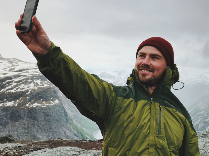 A man in outdoor gear taking a selfie with a smartphone in a mountainous landscape, illustrating dumb ways people ruin their lives.