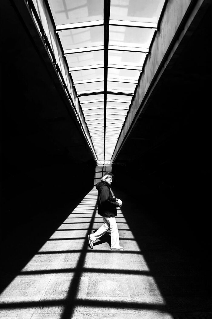 Black and white minimalist architecture, a person walking through a shadowy tunnel with light patterns from a skylight.