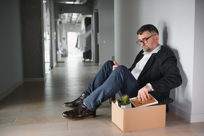 A man sitting on the floor with a box of belongings, representing ways people ruin their lives through job loss.