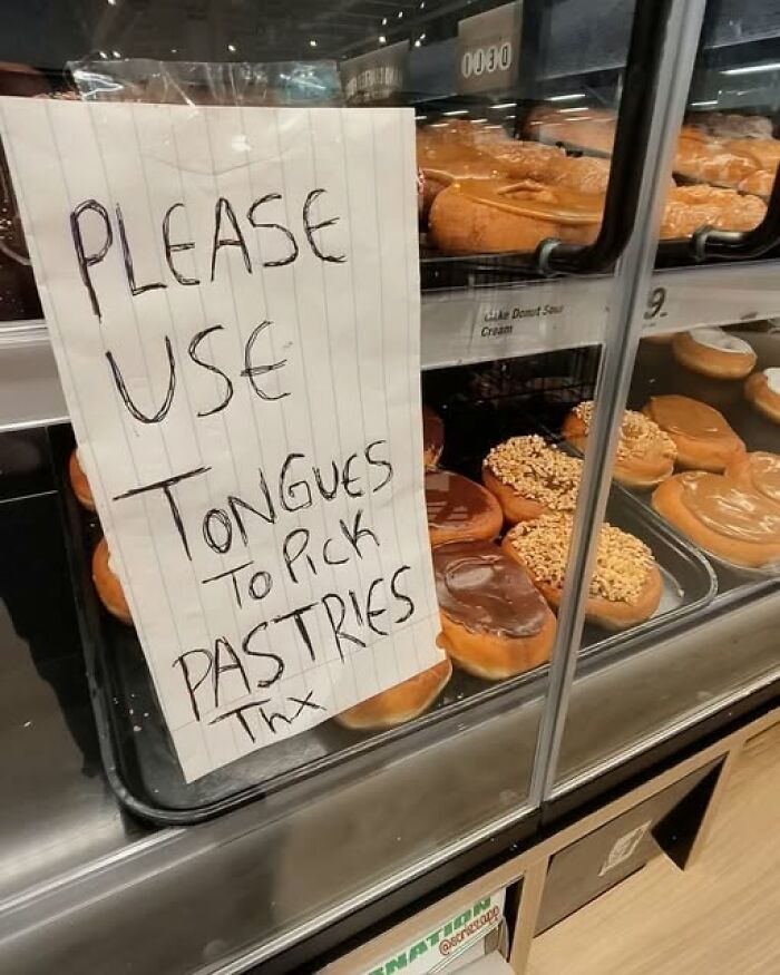 A silly sign in a bakery asking customers to use tongues to pick pastries, next to a display of funny donuts.
