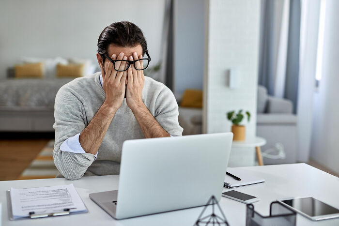 A man with glasses covering his face at a laptop, showing dumb ways people ruin their lives.