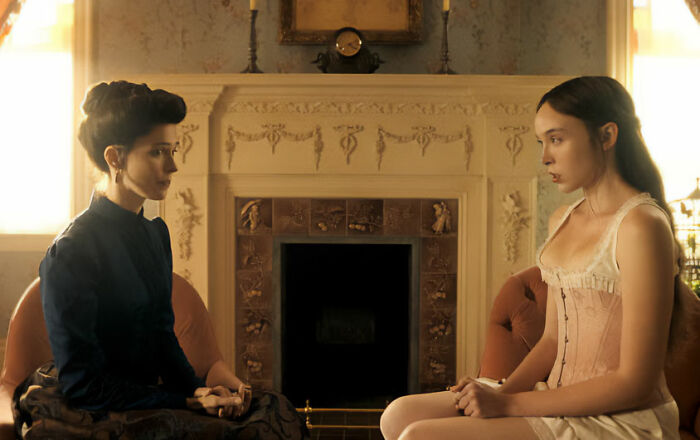 Two women in period attire sitting by a fireplace, one in a dark dress, the other in a corset, from the nepo baby's show.