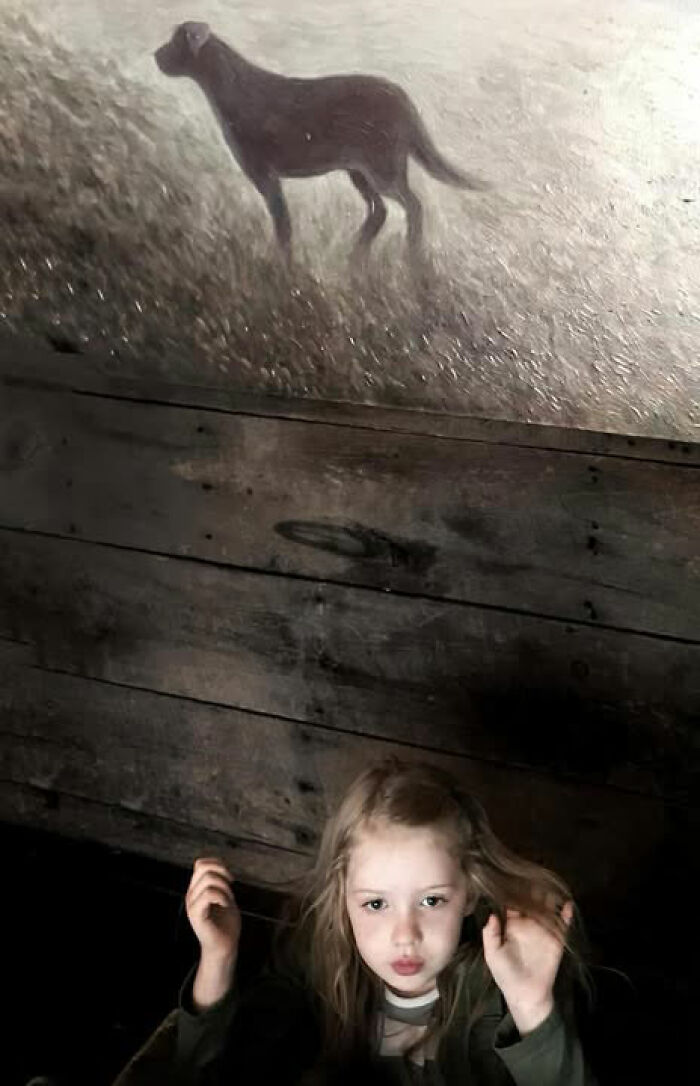 A girl looks up at a painting of a dog on a dark wooden ceiling, a street photo by Taylor Coble.