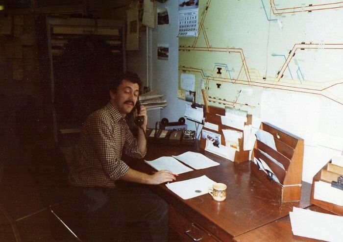 A man with a mustache talks on a retro tech phone in an office, surrounded by papers and a map, capturing the 80s weirdness.