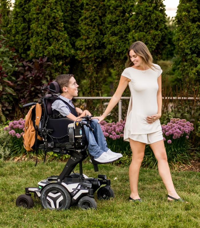 Pregnant TikTok star standing beside her husband in a wheelchair, both smiling in a garden, celebrating their pregnancy. Pregnant TikTok star standing beside her husband in a wheelchair, both smiling in a garden, celebrating their pregnancy.