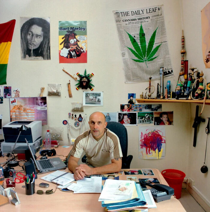 A man sits at his desk in an office filled with posters and paraphernalia, documenting the people and offices behind bureaucracy.