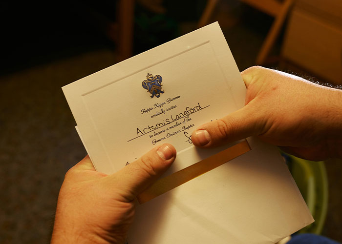 Hands holding an official invitation letter addressed to Artemis Langford, related to the sorority sisters case.