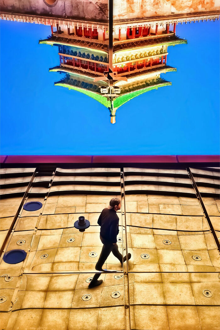 A unique perspective of a man walking on a reflective surface, with an inverted architectural building above, creating a minimalist scene.