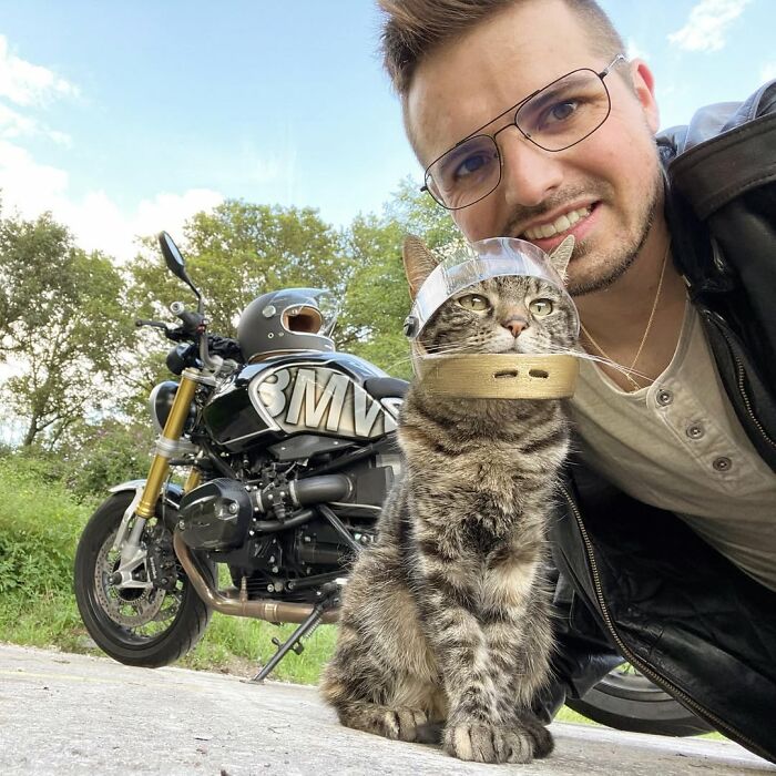 An owner and his cat, both wearing helmets, posing next to a motorcycle, ready for crazy adventures.