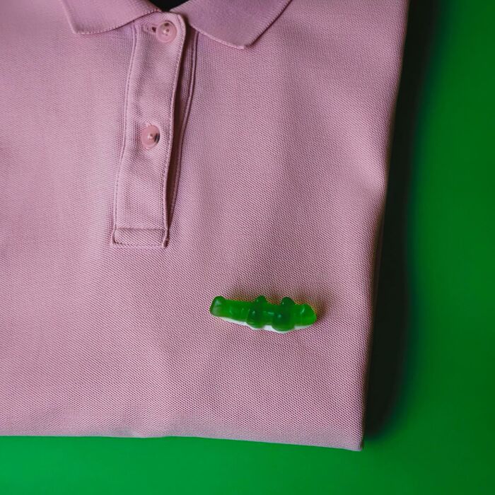 A green gummy alligator replaces the logo on a pink polo shirt in a playful illustration.