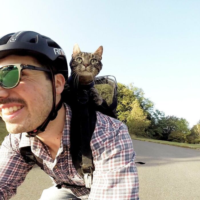 A man wearing a bike helmet and sunglasses, smiling as his cat rides in a basket on his back, on an adventure.
