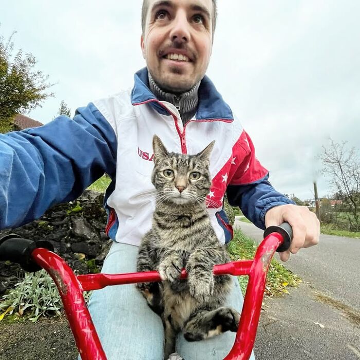 A man smiles while riding a red bike, his cat perched comfortably between his legs, embarking on new adventures.