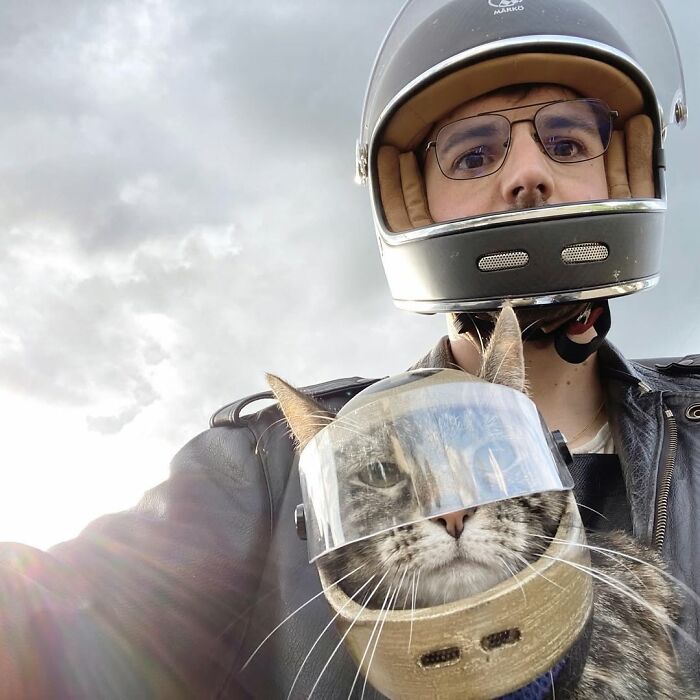 Owner and cat in matching helmets, ready for crazy adventures on a motorcycle under a cloudy sky.