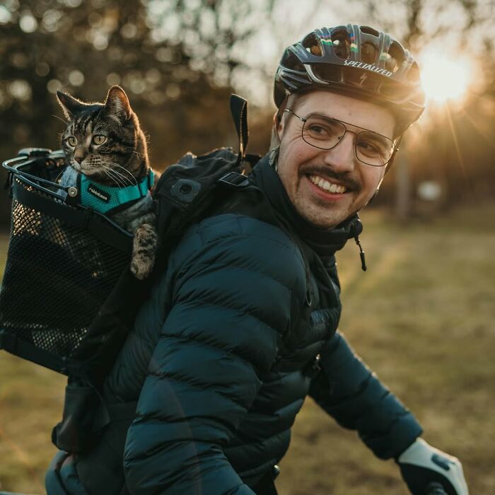 Owner bringing his cat everywhere, cycling with the cat in a basket on his back during sunset.