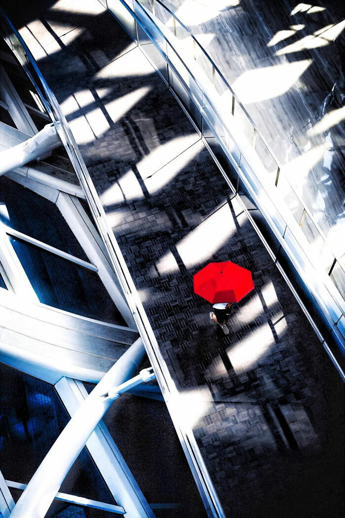 A photographer captures everyday architecture into a bold minimalist scene of a person with a bright red umbrella walking across a modern bridge.