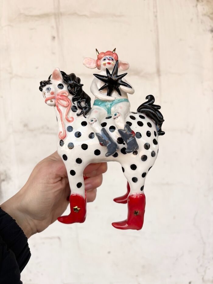 A hand holding a weird ceramic sculpture of a spotted horse with red boots and a character rider.