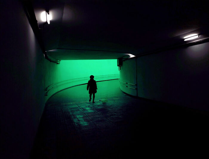 A photographer creates minimalist architecture, with a silhouetted person walking towards a bright green light in a dark tunnel.