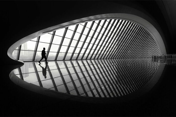 A lone person walks across a minimalist scene, with bold architecture reflecting on a polished floor.