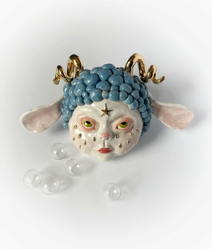 A finished weird ceramic sculpture showing a horned creature face with gold horns and blue hair.